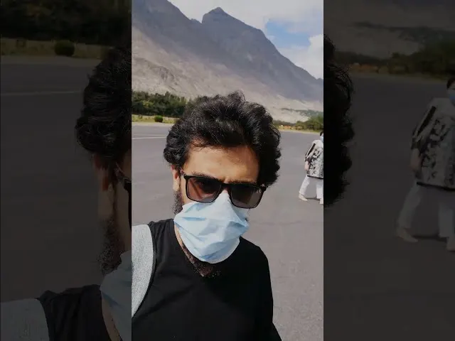 Man wearing sunglasses and blue mask at airport with mountain backdrop